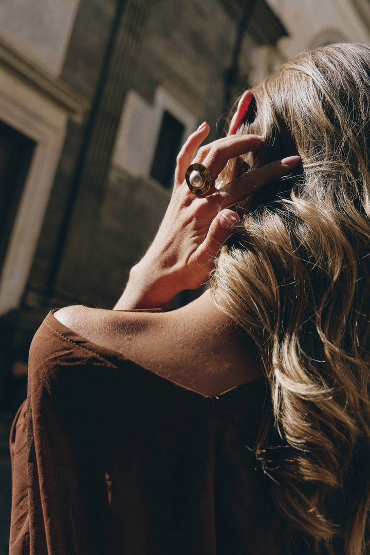 Anello Selene in metallo dorato con perla, indossato su una mano di donna con capelli mossi, vista di schiena, sfondo urbano.